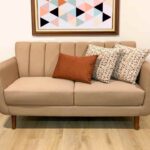Sofá moderno tapizado en tela suave color camel, con cojines decorativos y patas de madera, ideal para una sala pequeña, elegante y con diseño