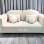 Sofá Leno de dos puestos en color claro, con diseño moderno y curvas suaves, patas de madera natural y cojines decorativos. Elegancia y confort para espacios acogedores