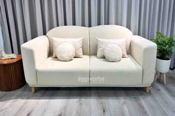 Sofá Leno de dos puestos en color claro, con diseño moderno y curvas suaves, patas de madera natural y cojines decorativos. Elegancia y confort para espacios acogedores