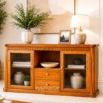 Bife clásico elaborado en cedro macizo con acabado natural, dos puertas en vidrio, dos cajones centrales y detalles tallados en la parte superior. Ambientado en una sala elegante de estilo clásico-contemporáneo con decoración cálida y sofisticada.