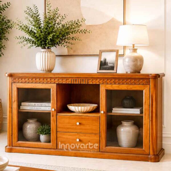 Bife clásico elaborado en cedro macizo con acabado natural, dos puertas en vidrio, dos cajones centrales y detalles tallados en la parte superior. Ambientado en una sala elegante de estilo clásico-contemporáneo con decoración cálida y sofisticada.